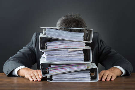 Frustrated businessman with lot of files on desk against gray background