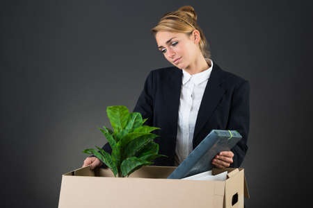 Sad young businesswoman packing her belongings at desk over gray backgroundの写真素材
