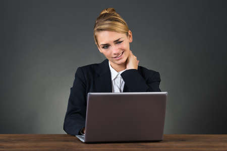 Young businesswoman suffering from neck pain while using laptop over gray backgroundの写真素材