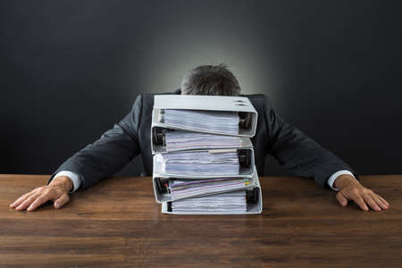 Frustrated businessman with lot of files on desk against gray backgroundの写真素材