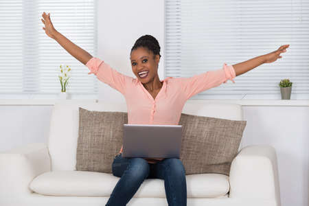 Portrait Of Excited African Woman Sitting On Sofa With Laptopの写真素材