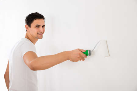 Young man using paint roller on white wall at homeの写真素材