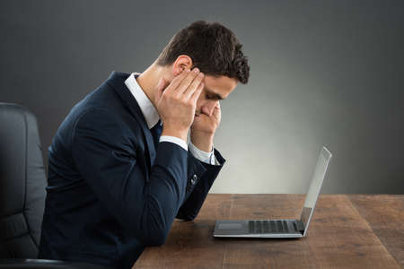 Businessman suffering from headache while working on laptop at desk against gray background