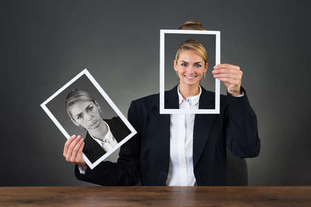 Young businesswoman holding photographs with different expressions at desk over gray backgroundの写真素材