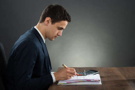 Side view of young businessman examining invoice at desk against gray backgroundの写真素材