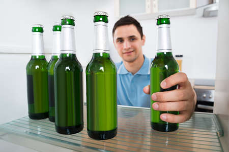 Smiling young man removing beer bottle from refrigerator at homeの写真素材