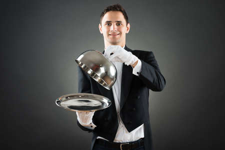 Portrait of happy waiter holding cloche over empty tray while standing against gray backgroundの写真素材