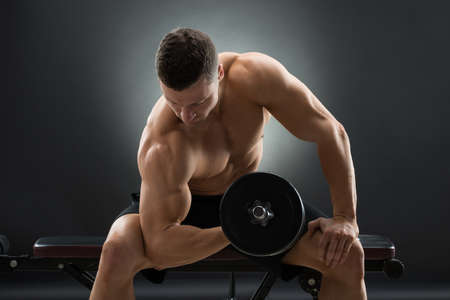 Muscular man exercising with dumbbells while sitting on chair against black background