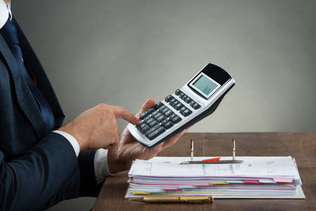 Midsection of businessman using calculator while examining invoice at desk against gray backgroundの写真素材