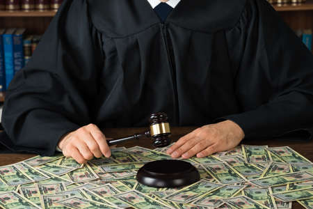 Midsection of corrupt judge hitting gavel with banknotes spread at desk in courtroom