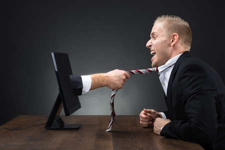 Businessman's hand emerging from computer monitor and pulling tie of executive at tableの写真素材