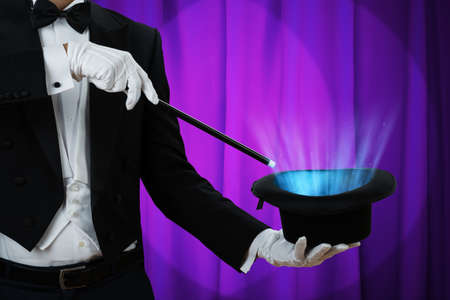 Midsection of magician holding magic wand over illuminated hat against purple curtain