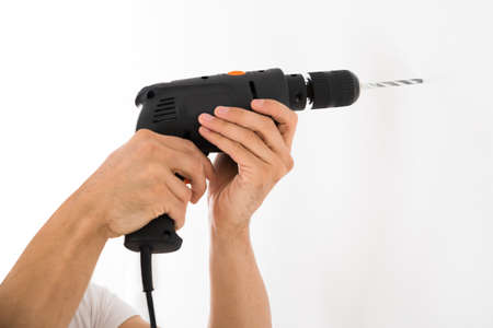 Cropped image of young man using power drill on white wall at home