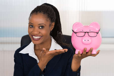 Young African Businesswoman Holding Piggybank At Office Deskの写真素材
