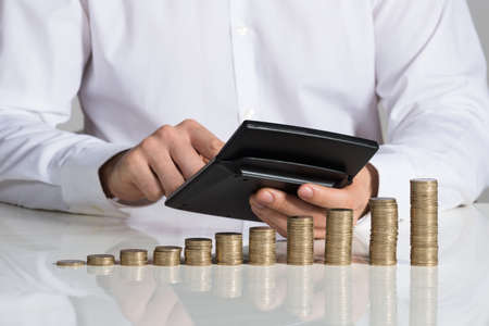 Midsection of businessman using calculator with stacked coins arranged at office deskの写真素材