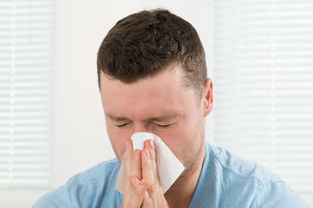 Closeup of mid adult man with cold blowing his nose at homeの写真素材