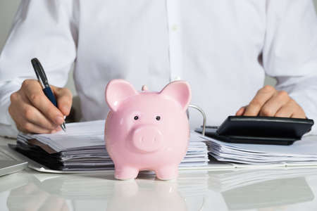 Midsection of businessman calculating invoice with piggybank at office deskの写真素材