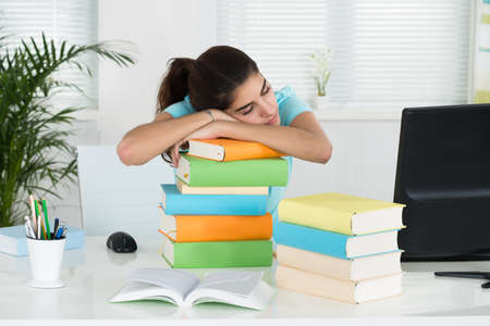 Young female student resting on stacked books at table in home office