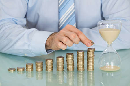 Close-up Of Businessman Holding Hourglass Near Stack Of Coins