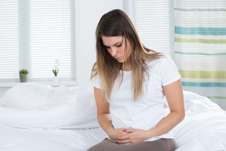 Unhappy Young Woman Suffering From Stomach Ache Sitting On Bedの写真素材