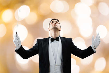 Young male music conductor looking up while holding baton against illuminated backgroundの写真素材