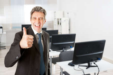 Portrait of mature businessman showing thumbs up sign by desk in officeの写真素材