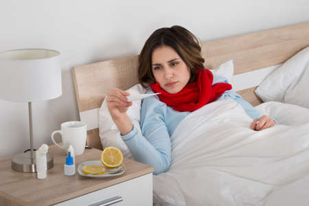 Sick Woman Lying On Bed Looking At Thermometer