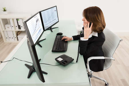 Young Female Stock Market Broker Working On Multiple Computers While Talking On Phone