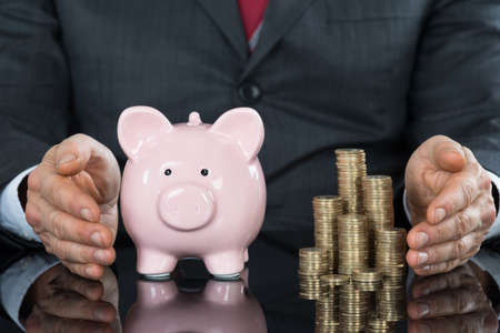 Close-up Of Businessman Hand Protecting Piggybank With Stack Of Coins