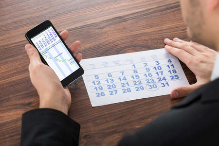 Close-up Of Businessman Holding Mobile Phone With Calendar On Desk