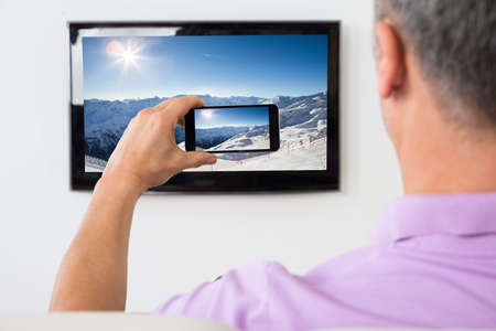 Mature Man With Smartphone Connected To A TV Watching Movie At Home