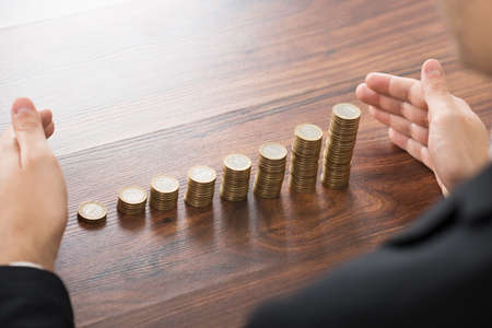 Close-up Of Businessman Hand Protecting Stacked Coins On Desk