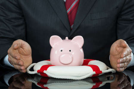Close-up Of Businessman Hand Protecting Piggybank With Lifebelt At Deskの写真素材