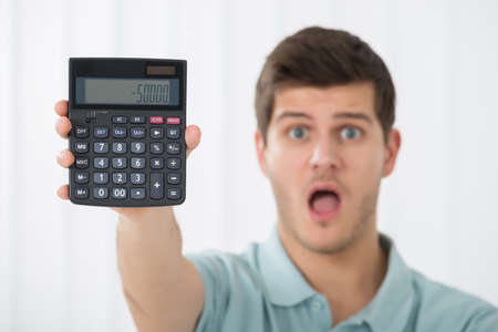 Shocked Young Man Holding Calculator At Homeの写真素材