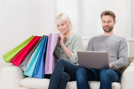 Young Couple Sitting With Shopping Bags And Laptop On Couch At Home