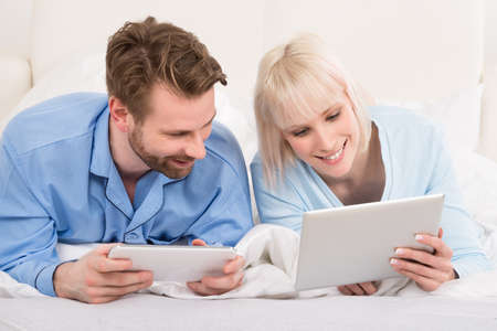 Young Happy Couple Lying In Bed With Digital Tablets In Bedroom At Homeの写真素材