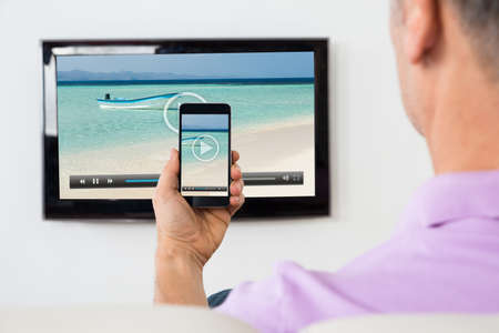 Mature Man With Smartphone Connected To A TV Watching Video At Home