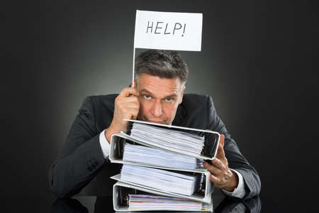 Sad Mature Businessman Holding Flag With Help Text Above Stack Of Folders On Deskの写真素材