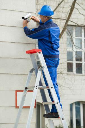 Mature Male Technician Standing On Stepladder Fitting CCTV Camera On Wallの写真素材