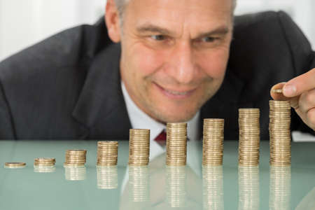 Mature Happy Businessman Placing Coin Over Stack Of Coins At Deskの写真素材