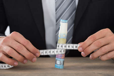 Close-up Of Businessman Hands Holding Banknote With Measure Tape On Deskの写真素材