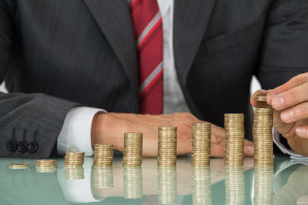 Close-up Of Businessman Hand Placing Coin Over Stack Of Coins At Deskの写真素材