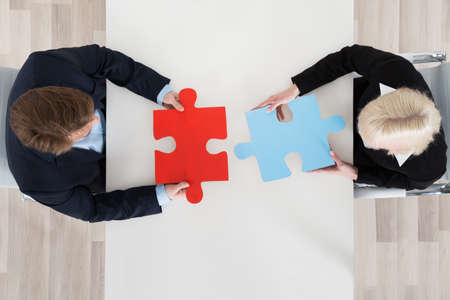 High Angle View Of Two Businesspeople Connecting Puzzle Pieces At Office Desk