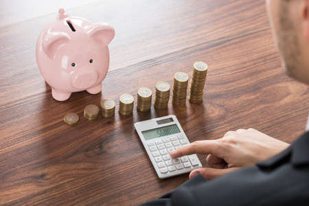 Close-up Of Businessman Using Calculator With Piggybank And Stacked Coins On Deskの写真素材
