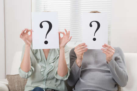 Couple Sitting On Sofa Holding Question Mark Sign In Front Of Faceの写真素材