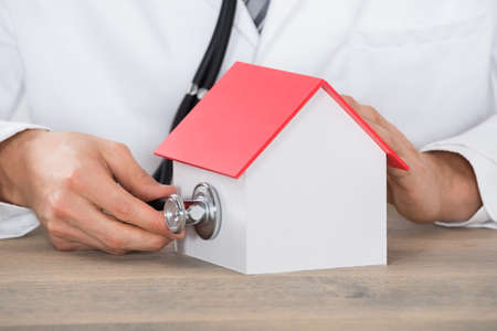 Close-up Of Doctor Hand Holding Medical Stethoscope On House Model At Wooden Deskの写真素材