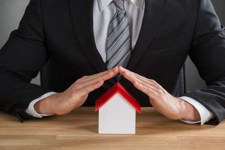 Close-up Of Businessman Hand Protecting House Model At Wooden Deskの写真素材