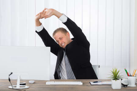 Young Businessman Stretching In Office With Computer On Deskの写真素材