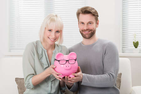 Happy Couple Sitting On Sofa Holding Piggybank At Homeの写真素材