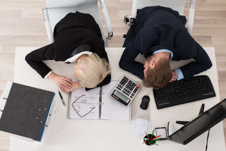 Two Tired Young Businesspeople Sleeping On Desk In Officeの写真素材